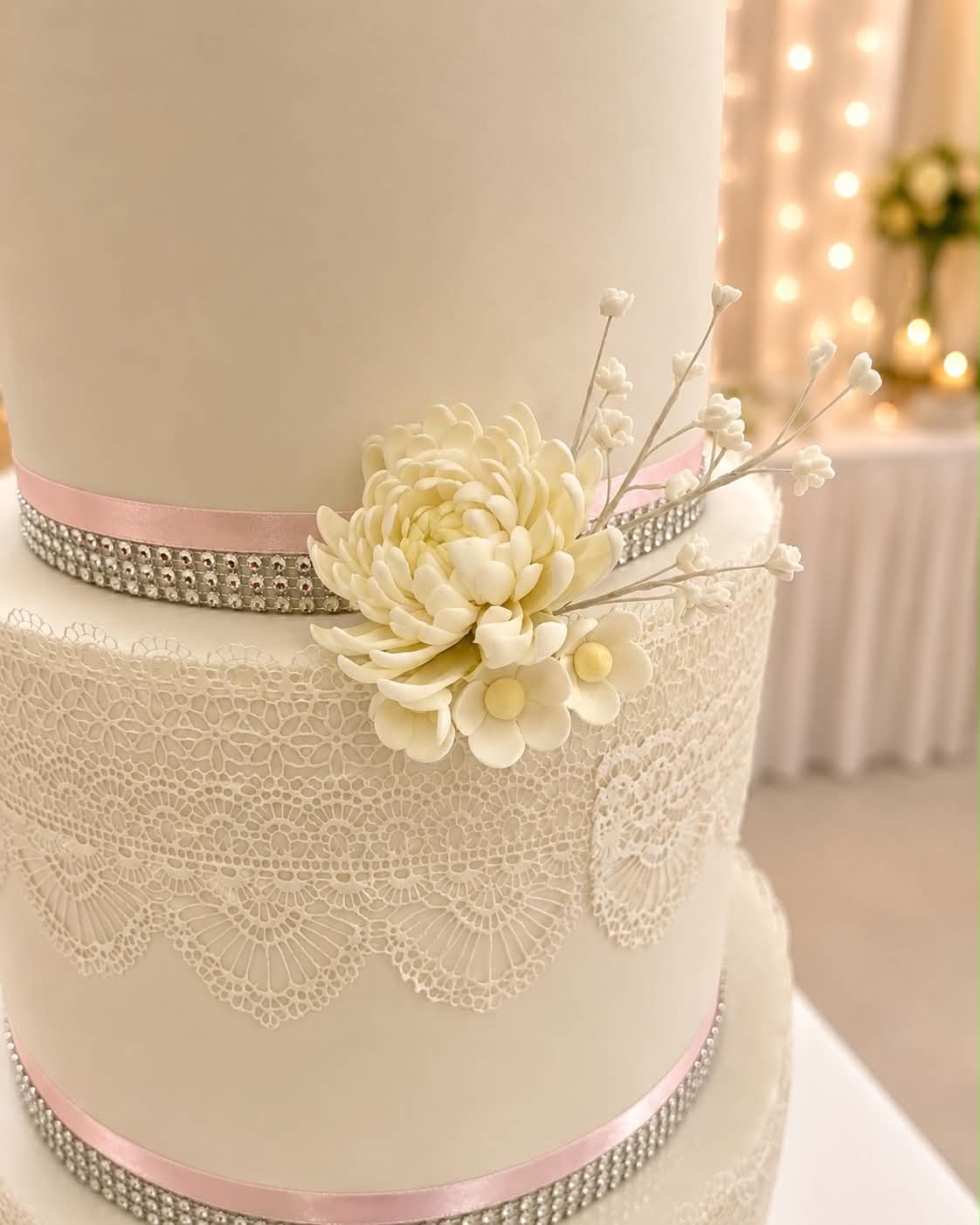 Close up of ivory fondant wedding cake tier with intricate lace pattern, diamante ribbon border, pink satin ribbon and a handcrafted ivory chrysanthemum and white blossom sugar flower arrangement